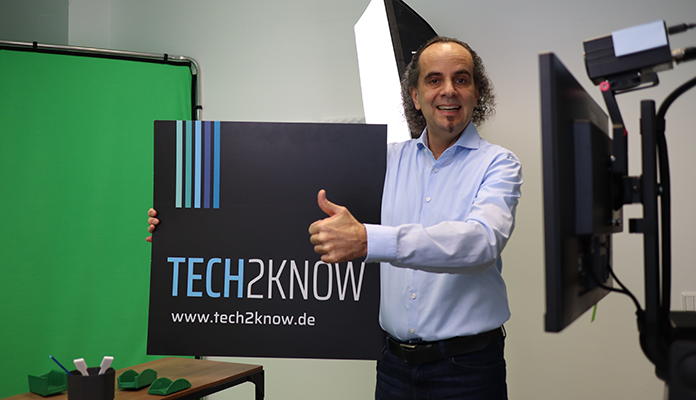 Aufnahme aus dem Tech2Know Video-Studio. Dr. Thornagel hält ein Schild mit dem Tech2Know-Logo hoch.
